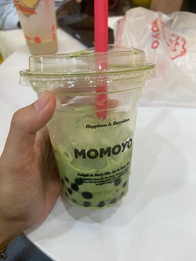 A close-up, slightly high-angle shot shows a hand holding a clear plastic cup filled with a green beverage and black tapioca pearls, commonly known as bubble tea. The cup has a clear plastic lid and a bright pink straw. The beverage has condensation on the outside of the cup, indicating it is cold. Text is printed on the cup, with the word "MOMOYO" prominently displayed in large, dark letters. Above "MOMOYO," smaller text reads "Happiness Is Everywhere," and below, additional text is partially visible, starting with "Delight in Every bite, Joy in Every." In the background, out of focus, is another clear plastic cup with a light-colored beverage and red accents, and a white plastic bag with red text and a graphic. The scene appears to be indoors, likely at a food establishment, with a white table surface in the foreground. There is no specific information about Kembang Harum, Indonesia present in the image.