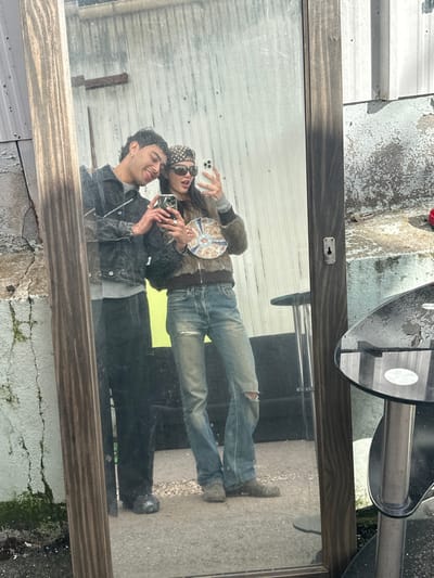 An outdoor mirror selfie taken in Neuilly-Plaisance, France, depicts two individuals. A male figure on the left, with dark wavy hair and earrings, smiles while embracing a female figure with his left arm around her shoulders. He wears a dark denim jacket over a light grey top and dark trousers, holding a dark smartphone in his right hand. The female figure on the right holds a light-colored smartphone in her left hand, which appears to be taking the photograph, and a decorative, possibly ceramic, plate in her right hand. She wears a brown patterned bandana, dark sunglasses, a brown fuzzy bomber jacket, a light grey long-sleeved top, distressed light blue jeans with rips at the knees, and dark studded shoes. Both are reflected in a large, upright mirror with a dark brown, possibly fluted, frame on the left and a plainer dark frame on the right.
The reflected background behind the individuals features a light-colored, worn corrugated metal or panel wall with visible peeling paint, topped by a darker corrugated surface. A horizontal metal beam is visible in the upper left reflected area. The reflected ground is a hard surface, likely asphalt or concrete. To the right of the mirror, outside the reflection, a black-topped, silver-pedestal table is visible. To the left of the mirror, an exterior wall shows significant weathering, peeling paint, and green moss or lichen growth. The scene suggests an outdoor, possibly industrial or utilitarian, setting.