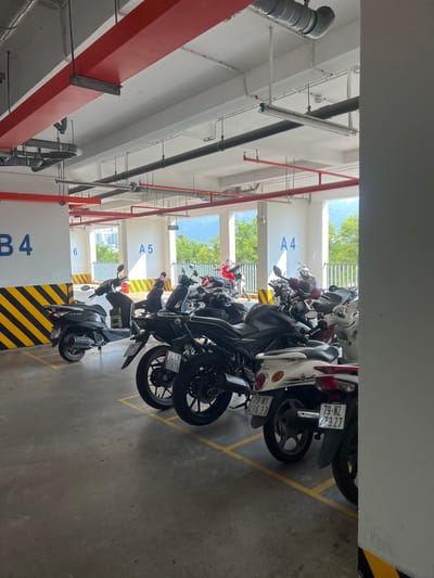 This image depicts a row of parked motorcycles in what appears to be an indoor parking garage. The parking spaces are marked with yellow lines and numbered, with visible signs displaying "A4," "A5," and "B4." The motorcycles vary in color and style. The ceiling of the garage has visible piping and support beams. Through the gaps between the parked motorcycles, trees and buildings can be seen, indicating an exterior view. The location is identified as the Nam Natural Heritage Area Trang, Vietnam.