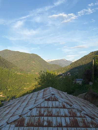 The image shows a landscape near Kveda Chkhutuneti, Georgia. The shot is angled, presenting a view of mountain ranges with a clear blue sky interspersed with light clouds. The mountains are covered with green vegetation, and a second mountain range in the distance appears to have snow-capped peaks. In the foreground, a corrugated metal roof is visible, occupying a significant portion of the right side of the frame. The structure associated with the roof is not entirely in view.