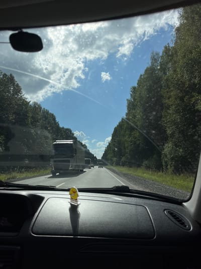 The image shows a view from inside a car driving on a highway in Uspenskii, Russia. A large semi-truck is visible in the distance on the right lane of the highway, followed by other vehicles. The highway is lined with dense green trees on both sides, and the sky is blue with scattered white clouds. The dashboard of the car is in the foreground, with a small yellow figurine of a chick placed on it. The overall scene depicts a sunny day on a road trip.