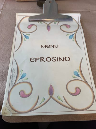 The image displays a slightly rotated close-up of a menu cover attached to a metal clipboard, resting on a textured surface, possibly a table. No people are visible.
The menu cover is a light cream or pale yellow color, adorned with decorative swirling borders in brownish-gold tones. Within these swirls are colorful, leaf-like or teardrop shapes in shades of pink, purple, light blue, and light green, adding a vibrant touch to the design.
Vertically oriented in the center, the word "MENU" is printed in bold, capitalized brown letters. To its right, also vertically oriented and in bold, capitalized brown letters, is the name "EFROSINO". In the bottom right corner of the menu cover, a handwritten signature "M. Βασιλιριου" is visible, followed by the year "2018", suggesting an artist's signature and the year the design was created.
The clipboard's metal clamp is visible on the left side, securing the menu. The menu cover shows slight signs of wear, with some minor creases and a slightly bent corner at the bottom right. The background is a soft, out-of-focus beige or brown. The overall impression is that of a restaurant menu from Kastraki, Greece, with a handcrafted artistic design.