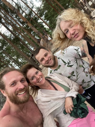 A selfie taken outdoors on a sandy area with tall pine trees in the background. Four people are visible: a shirtless man with a beard and blue eyes in the foreground on the left, smiling; a woman with shoulder-length brown hair, wearing a green bikini top and a beige striped shirt, smiling in the center; a man with short brown hair and a beard, wearing a white t-shirt with a floral print, smiling in the upper center; and a woman with long blonde curly hair and light-colored eyes, winking, in the upper right. A small brown dog is partially visible on the far right. The background shows the sandy ground, tree trunks, and foliage of a pine forest. Structures, possibly wooden, are visible behind the people amongst the trees. The overall atmosphere appears casual and friendly. This scene is likely taking place in Luzhki, Russia, suggested by the dense pine forest typical of the region.