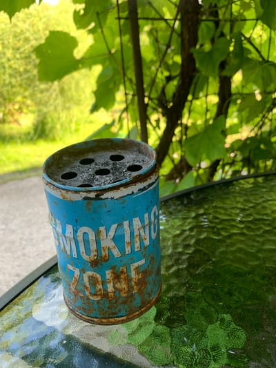 The image depicts an old, rusted blue metal ashtray on a textured green glass table. The ashtray has white lettering that reads "SMOKING ZONE" and has several holes at the top, filled with ash. In the background, there are green leaves and branches of trees, with a blurred outdoor setting suggesting a park or garden area. The lighting indicates it is daytime, with sunlight filtering through the foliage. The scene appears to be in Silmala, Latvia, implying an outdoor public space where smoking is permitted. No people are visible in the image.