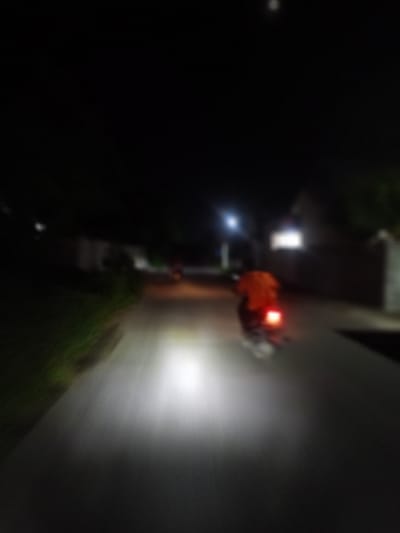 A blurred image depicts a night scene in Geulanggang Baro, Indonesia, showing a person on a motorbike driving on a road. The motorbike's headlight illuminates the road ahead. Streetlights are visible, and buildings line the road.