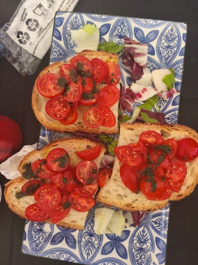 This image shows a close-up overhead view of two pieces of bruschetta, topped with sliced cherry tomatoes and a green herb dressing, placed on a blue and white patterned plate. A small salad of mixed greens and radicchio is also on the plate. To the left of the plate, a red plastic bottle with some white labeling and a plastic bag with Italian text and recycling symbols are visible. The food appears to be on a dark, textured surface, possibly a table. The presence of a red bottle with recycling symbols and Italian text suggests the location is likely in Italy. Specifically, a red bottle with "RICICLABILE" (recyclable) printed on it, and a plastic bag with "IMBALLAGGIO ESTERNO" (external packaging) and Italian text, further confirms an Italian context. The specific mention of "San Salvo, Italy" is not directly observable from the visual elements in the image itself, but the packaging and language strongly indicate Italy.
