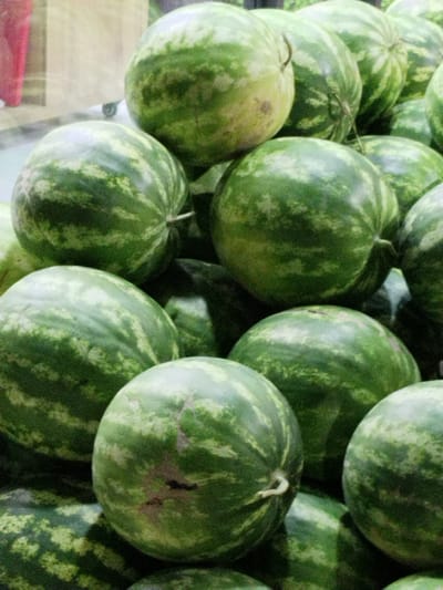 The image shows a pile of watermelons in what appears to be a market stall. The watermelons are green with darker green stripes and vary in size and shape. The lighting is somewhat dim, casting shadows on the fruits. In the background, a blurry section of a wooden surface and some reflective material are visible, suggesting an indoor or covered outdoor setting. There are no people or visible interactions in the image. The scene does not provide any specific clues about its location within the city of Jizzakh, Uzbekistan.