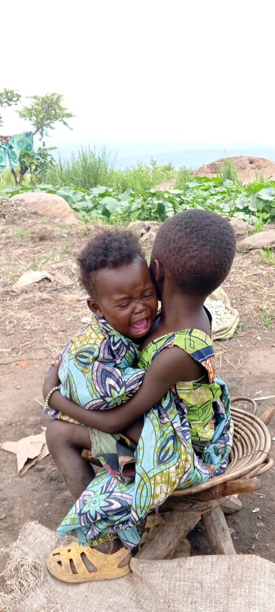 Two children are outside in a rural area. The smaller child is crying while being held by the larger child. The children are wearing matching patterned clothing. The crying child is wearing a bracelet and a yellow shoe is visible. A woven basket and wooden supports are on the ground next to them. The background shows green vegetation and a distant landscape.