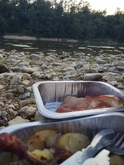 The image depicts a close-up view of two foil pans filled with food, placed on a rocky riverbank. In the foreground, a pan contains what appears to be roasted potatoes and some sort of sausage or meat. Further back, another pan holds raw meat, possibly pork, with some fat visible. The riverbank is covered in smooth, grey and beige stones of varying sizes, with sparse green grass growing between them. The water of a river is visible behind the pans, and on the far side of the river, a dense forest line forms the horizon under a bright sky. The image composition suggests an outdoor meal setting, likely a picnic or camping trip. The location is identified as Boé, France, though no specific Boé landmarks are visible in the image.