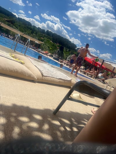 The image captures a scene at a poolside location, likely a resort or public pool, on a sunny day. The foreground is dominated by a textured, light-colored tiled surface, with shadows cast by foliage and a black chair. In the mid-ground, a swimming pool with clear blue water is visible, featuring metal ladders for entry. Several people are present, some lounging on sunbeds and others in the pool. One man in patterned swim shorts is walking away from the camera, holding a green ball. The background shows a lush, tree-covered hillside with scattered buildings, indicative of a location near a town or city, possibly Delchevo, North Macedonia, given the context. The sky is bright blue with scattered white clouds.