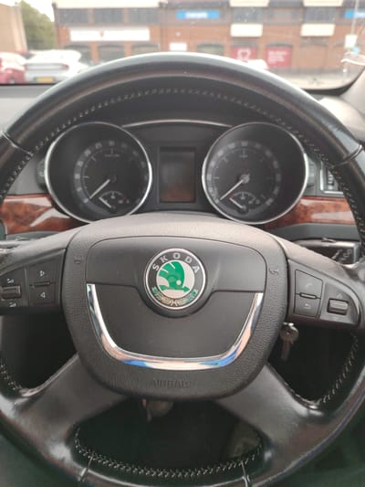 The image depicts the interior perspective from a stationary Skoda vehicle, looking forward through the windshield. In the immediate foreground, a black leather steering wheel with visible stitching dominates the lower half of the frame. The central hub of the steering wheel displays the green Skoda emblem. Integrated controls are present on both the left and right spokes; the left features arrow buttons and a mode selector, while the right includes phone call management buttons and additional arrow selectors. A small, metallic key is seen dangling from the right side of the steering column area.
Behind the steering wheel, a segment of the instrument cluster is visible, featuring a tachometer on the left and a speedometer on the right, both indicating zero. A dark, possibly faux wood, trim panel extends across the passenger side of the dashboard.
Through the windshield, an urban environment is visible under an overcast sky. Several vehicles are parked or driving in the distance; a red car is partially visible on the far left, and a white car with a yellow UK license plate is positioned further back in the center-left. The primary structure in the background is a reddish-brown brick building with multiple windows and white signage. Visible details on the building include a blue sign on the far left, a prominent red heart logo on a white background above a doorway, and a red sign to its right with partial text "ALEXANDRA" and possibly "ALEXANDRA R..." or "ALEXANDRA ROAD" below it. Another blue sign is visible further right. Green foliage from trees is visible on the far left.
Based on the visible signage, including "ALEXANDRA" and the distinctive red heart logo, the location is identified as Alexandra House, situated on Alexandra Road in Peterborough, United Kingdom. The vehicle is parked facing this building.