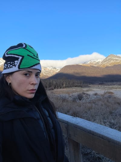 A woman wearing a green, black, and white beanie and a black jacket is positioned in front of a landscape. She is likely on a wooden structure. In the background, there is a mountain range partially covered by low-lying clouds. The sky is blue, and the vegetation appears to be dry grasses or brush. The location is unknown.
