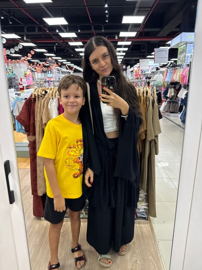 In a retail store, possibly in Phu Thuy, Vietnam, a woman and a young boy stand together, reflected in a large mirror. The woman, with long dark hair, wears a white crop top and a long black open-front garment, holding a smartphone in her right hand. The boy, to her left, smiles at the camera wearing a bright yellow t-shirt with a cartoon graphic and black shorts. They are surrounded by racks of clothing for sale, with visible signage in a foreign language in the background. The lighting suggests an indoor, commercial environment.