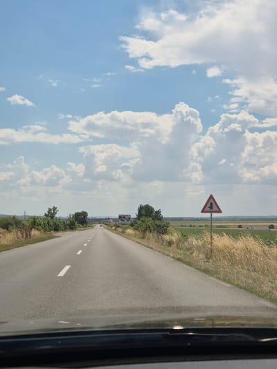 This media file shows a view from a vehicle driving on a paved road. The road has white dashed lines indicating lanes and a solid white line on the right edge. The surrounding landscape consists of dry, golden grasses on the right and green vegetation and trees on the left. In the distance, there are hints of agricultural fields. The sky is a clear blue with large, puffy white clouds.
A triangular traffic sign with a red border is visible on the left side of the road, indicating a junction ahead where traffic from the left must yield or turn. Further down the road, a billboard can be seen on the right. A few other vehicles are also visible in the distance on the road.
The time of day appears to be daytime, with bright sunlight casting shadows, and the weather seems pleasant and clear. The location is identified as Veselinovo, Bulgaria. The image evokes a sense of travel and the open road under a dynamic sky. There is no visible text other than on the distant billboard.