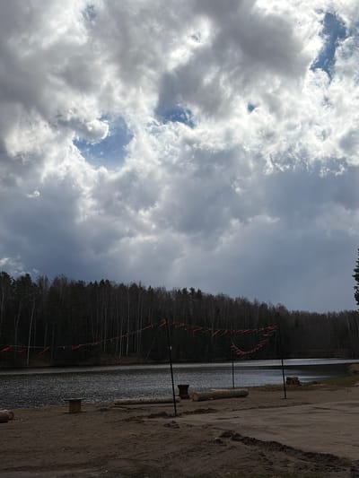 This outdoor landscape scene, set in Mishkino, Russia, features a body of water, a forested shoreline, and a partly cloudy sky. The upper portion of the image is filled with a dynamic sky, characterized by a mix of bright white and gray cumulus clouds. Several patches of clear blue sky are visible between the clouds, indicating varying light conditions, likely daytime.

A dense forest, appearing to consist mainly of bare deciduous trees, lines the far bank of the expansive body of water, which occupies the middle ground. The water's surface shows ripples and reflects the light.

In the foreground, a wide, unpaved dirt or sandy bank stretches alongside the water. Several dark-colored utility poles or posts are erected along this bank and extend into the water. A line, possibly a rope or cable, is strung between some of these poles, from which numerous small, bright red flag-like markers or buoys are suspended over the water. These markers suggest a designated area or boundary, potentially for a water event, a swimming zone, or to mark an underwater feature.

Two large, debarked wooden logs lie horizontally on the dirt bank in the foreground, and a smaller wooden block is visible closer to the water's edge. There are no people visible in the image, and no specific activity is taking place, though the presence of the marked line implies preparation for future events. No visible text is present.