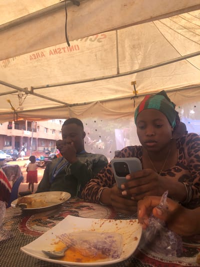 Two individuals are seated at a table, likely under a tent, in Nteje, Nigeria. The woman, wearing a leopard print top and a headwrap, holds a mobile phone and a plastic bag. A man sits across from her, wearing a green top and a lanyard. The table has plates of food, some partially consumed. The background reveals a street scene with buildings and other people.
