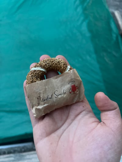A hand holds a freshly baked simit, a Turkish bagel covered in sesame seeds. The simit is partially wrapped in brown paper, which is printed with the words "Istanbul Simidi" and a small illustration of a tea glass. The simit is resting on the palm of the hand, with the fingers curled around it. The background is a teal-colored fabric. The image appears to have been taken outdoors. The setting is not specified to be in the city of Ochamchyra, Abkhazia.