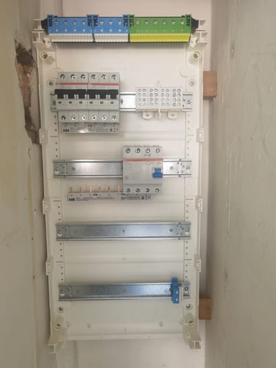 The image shows a white electrical distribution board mounted on a wall. The board contains several components, including circuit breakers, a distribution block, and DIN rail mounts. The board is unfinished and appears to be in the process of installation, with some wiring exposed. The wall behind the board is a light color and appears to be concrete or plaster. A wooden block is visible behind the right side of the board, likely used for mounting or support. The scene is indoors, possibly in a residential or commercial building. No people are visible in the image. The location of Karlsruhe, Germany, is not directly evident from the image itself but is provided as contextual information.