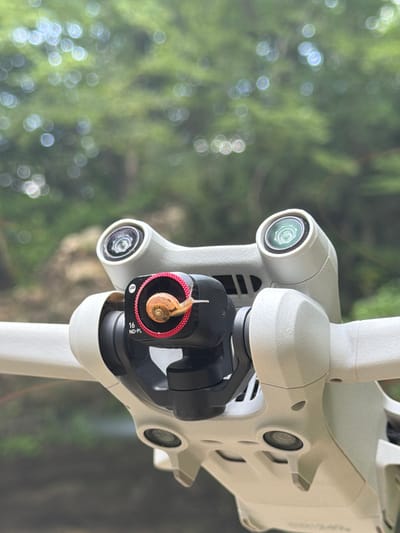 A close-up, slightly angled shot captures the camera gimbal of a white drone, focusing on a small snail perched on a lens filter. The drone's body, made of light-colored plastic, is prominently displayed, with its camera module and arms visible. The background is a soft, blurred green, suggesting an outdoor, natural environment, possibly with trees and foliage. The overall impression is one of a unique encounter between technology and nature, highlighting the snail's unexpected presence on the drone's equipment. No specific location in Kinchkhaperdi, Georgia is identifiable from the visual information.