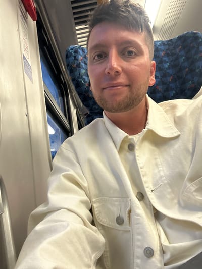 The image is a selfie taken on a train, showing a man wearing a white jacket. The interior of the train is visible, with a blue patterned seat behind the man. The window is partially visible, with the train's interior trim and a safety instruction label appearing in the foreground. The location is unknown, as the city of Balabanovo, Russia is not discernable from the visual elements present in the image. There is no verifiable evidence to support this location.