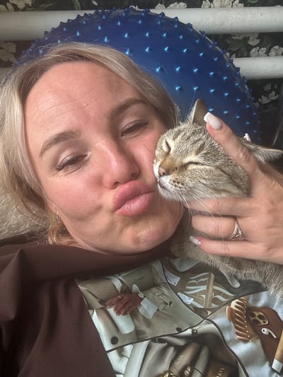 The media file shows a close-up, sideways selfie of a woman and a cat. The woman has blonde hair and is wearing makeup, including false eyelashes and lipstick. She is wearing a brown top. A tabby cat is nuzzling against her face. The woman's left hand is visible, resting on the cat's head. She is wearing a ring on her left ring finger.
The background includes a blue spiky massage ball and some decorative items. A patterned scarf is visible, featuring what appears to be a fashion-themed print with text that includes "ZARA" and "ILIA". There are also some cosmetic items like lipstick and mascara tubes scattered around. The overall impression is one of a casual, intimate moment between a person and their pet, possibly at home. The lighting suggests it is daytime. The image appears to be taken in Pozdyshki, Russia.