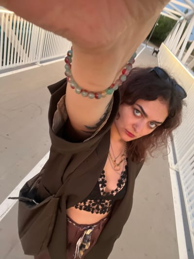 The image depicts a selfie-style, low-angle shot of an individual on a modern pedestrian bridge or elevated walkway, identified as being in Alcàsser, Spain. The subject, a person with wavy, reddish-brown hair, wears sunglasses pushed onto their head, a dark brown open jacket, and a black lace bralette. Multiple metallic necklaces with various pendants are visible around the neck, and a multi-colored beaded bracelet (red, green, light blue) is prominent on the right wrist, which is extended towards the camera. A dark tattoo is partially visible on the subject's inner right arm. The subject's gaze is directed towards the camera, and red lipstick is visible. The background features a light-colored paved walkway flanked by white railings constructed with vertical slats. In the distant background on the right, a small, dark-colored dog is visible walking on the path, alongside a street lamp. Beyond the bridge structure, green foliage and other structures are discernable. The scene is illuminated by natural light, creating some visible shadows.