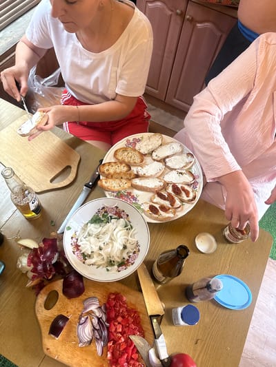 In Усть-Мана, Russia, two individuals are preparing food at a wooden table. A woman in a white t-shirt and red shorts is spreading a white substance on slices of bread with a knife. Next to her is a cutting board with more bread slices and a plastic bag. In the center of the table, a plate holds several prepared bread slices, some with the white spread and others topped with sun-dried tomatoes. A bowl of sliced white onions with herbs is also on the table, along with chopped red onions and tomatoes on a cutting board. Several bottles, a small jar, and a blue lid are scattered around the workspace. The environment appears to be a kitchen or dining area, with wooden cabinets visible in the background.