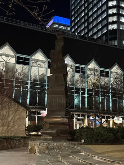 A bronze monument with stacked cubes and book-shaped pillars sits in front of a modern building with triangular roof peaks and gridded windows. The statue is inscribed with poetry. In the background, a tall skyscraper gleams with blue and white lights. Bare tree branches frame the scene, adding a touch of winter atmosphere to the urban setting.