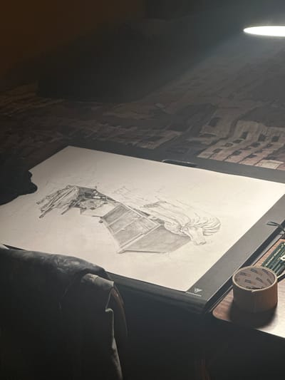 A drawing of a bureau with clothes strewn underneath is on a drawing board. A roll of tape and pencils rest on the board next to the drawing. The drawing is next to a brick wall with a bright light. The location is Montreuil, France.