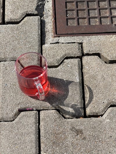 The scene depicts an outdoor paved area in Murzasichle, Poland, under direct sunlight. The ground is constructed from interlocking, light grey concrete pavers with a granular texture. In the upper right section, a rusty, dark brown metal drain cover with a square grid pattern is partially visible. A clear glass mug containing a translucent red liquid rests on the pavers in the central-left area; direct sunlight causes the liquid to cast a reddish illumination onto the adjacent paver surface. Below and slightly to the left of the mug, a dark-colored cylindrical object, identified as a "Winston" cigarette or vape stick by branding, lies horizontally on a paver block. Strong shadows are cast by the mug, the drain cover, and the paver edges, indicating a high sun angle. No individuals are present in the image.