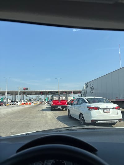 The image shows a view from inside a vehicle, likely a car, at a toll booth. The location is inferred to be near Santa María Moyotzingo, Mexico, based on the license plate of the white Nissan Versa in front (YSC-582-A), which indicates the state of Puebla. Other vehicles are visible, including a large white truck and a red truck. The toll booths are overhead, with lights indicating which lanes are open or closed. The sky is blue and clear. The steering wheel of the photographer's vehicle is visible in the foreground. The scene depicts a typical traffic queue at a toll plaza on a sunny day. There are no visible people or interactions.