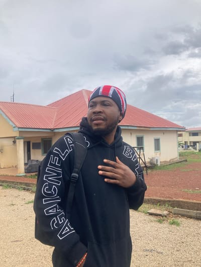 The image, oriented correctly, displays a young man of color posing outdoors in what appears to be Bisichi, Nigeria. He is positioned slightly off-center to the left, looking directly at the camera with a subtle, friendly smile. He has a short beard and mustache.
The man is dressed in a black hooded sweatshirt (hoodie) featuring prominent white block letters on both sleeves, clearly spelling out "BALENCIAGA" multiple times. Over his head, he wears a knitted beanie adorned with red, white, and blue stripes, resembling the design of the Union Jack flag. A black strap from a backpack is visible over his left shoulder, and his right hand is casually placed over his chest.
Behind him stands a building with a distinctive reddish-brown corrugated metal roof and cream or light yellow-colored walls. Several dark-framed windows are visible on the building's facade, along with an outdoor air conditioning unit. The ground in the foreground consists of a mix of gravel and reddish-brown earth, with patches of sparse green vegetation. Another building can be partially seen in the distance behind the main structure.
The sky is overcast with soft grey clouds, indicating a cloudy daytime setting. The overall atmosphere is casual and relaxed.