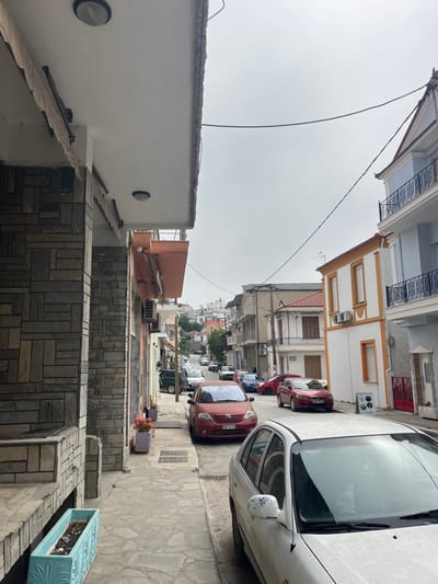 The image depicts a street scene in Limenaria, Greece, located at latitude 40.627 and longitude 24.575. The view is from the sidewalk, looking down a narrow street lined with buildings on both sides. The buildings are primarily two-story, with white stucco walls and terracotta tile roofs. The street surface is paved with gray stone. There are multiple parked cars lining the street, including a red sedan, a white sedan, and a silver hatchback. In the foreground, a person is seated in the driver's seat of a white sedan, looking out the front windshield. The sky is overcast with a pale gray color. The scene is quiet, with no visible people or activity beyond the driver in the foreground.