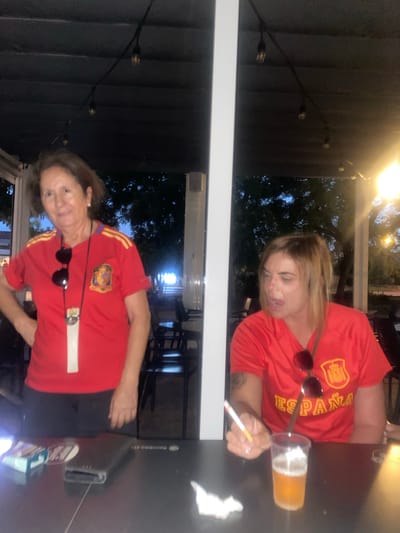 Two women, likely supporters of the Spanish national team, are depicted in a dimly lit outdoor setting. They are both wearing red shirts with the Spanish flag emblem and the word "España" visible on one of them. The woman on the left is older, with short brown hair, and is wearing sunglasses around her neck. The woman on the right has blonde hair and is holding a lit cigarette. In front of them on a dark table are a beer in a plastic cup, a pack of cigarettes, and a mobile phone. The background suggests a relaxed evening environment, possibly a patio or terrace area with string lights overhead and trees visible in the distance. The scene is indicative of a casual gathering to watch a sporting event or celebrate.