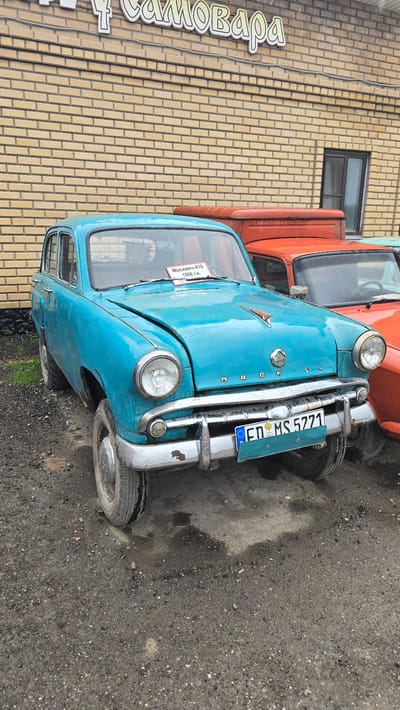 In Bol'shoi Mashlyak, Russia, a turquoise vintage Moskvich-410 car from 1958 is parked outdoors on a gravel lot in front of a light brown brick building. The building has a sign with Cyrillic text that reads "САМОВАРА" and a dark-framed window. The car has a chrome bumper, circular headlights, and a hood ornament. A paper sign on the windshield reads "Москвич-410 1958 г.в." and the license plate is "ED MS 5771". Another orange vehicle is partially visible behind the Moskvich. The ground is covered in gravel and there are a few dark wet spots, possibly from recent rain.