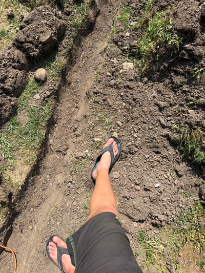 The image shows a person's legs and feet, clad in black flip-flops and dark shorts, standing on disturbed soil. The ground is uneven, with a visible trench or furrow dug into it, surrounded by clumps of dirt and scattered grass. A round, light-colored stone is visible to the left of the trench. The overall environment appears to be an outdoor, possibly rural or garden setting, with sunlight casting shadows. The location is identified as Bašť, Czechia.