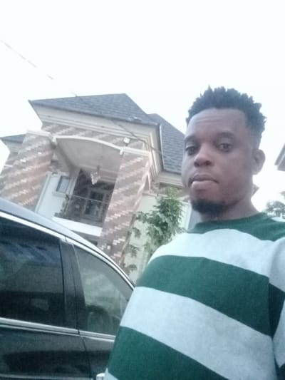 A man poses for a selfie outdoors with a vehicle and building in the background. The building is two-story with a gray roof and a porch. The man has dark hair and a beard, and he's wearing a green and white striped long-sleeved shirt. The car is dark-colored. The overall lighting suggests daytime, although somewhat overcast.