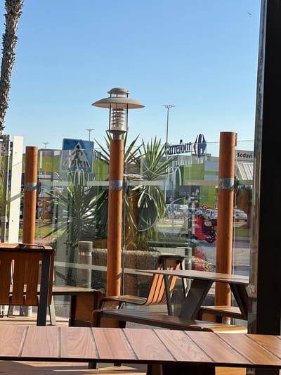 The image depicts an outdoor seating area with wooden tables and benches, likely part of a fast-food establishment, with the "Carrefour" supermarket visible in the background. The scene is set in the city of Sedaví, Spain. A street with vehicles, a pedestrian crossing sign, and a red scooter are visible through a glass partition. A palm tree is on the left side of the frame. The sky is clear and blue.