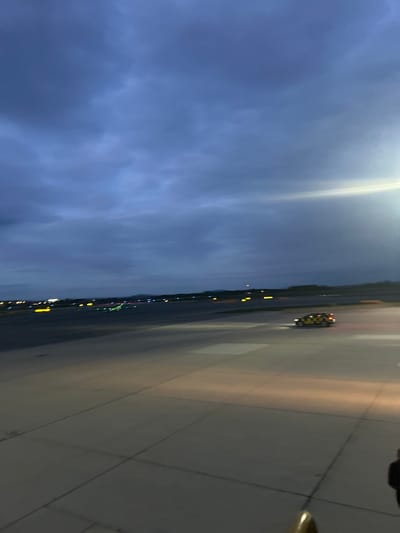The image, taken at Schwechat, Austria, depicts an airport scene at dusk or night. The perspective looks across a wide tarmac or taxiway, with the sky occupying the upper portion and the ground the lower.
The sky is dark, exhibiting shades of deep blue and purple, with visible cloud formations. A very bright, elongated light source is prominently situated in the upper left of the frame, casting a strong illumination. The ground, likely concrete or asphalt, covers the bottom half of the image, appearing expansive and marked by visible lines. It reflects the artificial light, indicating a smooth surface.
Along the distant horizon line where the sky meets the ground, a series of smaller, colorful lights—including yellow, red, green, and blue—are visible, characteristic of airport runway or taxiway edge lighting, extending into the distance. On the tarmac, in the middle-right section of the image, there is a distinct yellow and black checkered vehicle, which appears to be an airport "Follow Me" car or ground support vehicle, with its lights illuminated.
In the immediate bottom right corner, a dark, irregularly shaped object, possibly part of a window frame or the camera's immediate surroundings, along with a small, partially visible golden-colored element, are present. The scene suggests ongoing airport operations during the evening or nighttime. No visible text is discernible in the image.