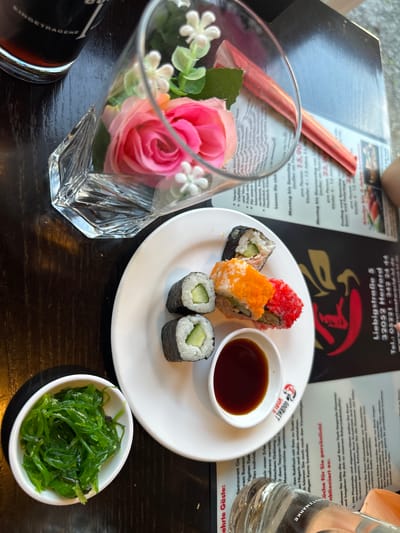 The image shows a table setting at "Gourmet World Herford," located at Liebigstraße 5, 32052 Herford, Germany. The table is dark wood, and a menu is visible. On the table, there is a white plate with several pieces of sushi, including cucumber rolls and a roll topped with orange and red roe. A small dish of soy sauce is next to the sushi. To the left of the plate is a small white bowl filled with green seaweed salad. In the background, a glass vase with artificial pink and white flowers is placed on top of the menu. Part of a glass of dark liquid, likely a beverage, is visible in the upper left corner.