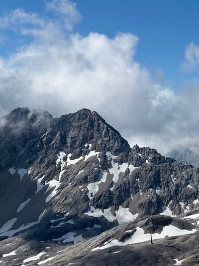 The image shows a jagged, rocky mountain range with patches of snow clinging to its slopes. The sky above is a mixture of blue with scattered white clouds. The terrain is rugged and appears to be at a high altitude. A single, tall, dark-colored pole-like structure with a square base is visible on a lower, snow-dusted ridge in the foreground. The mountain slopes are characterized by a mix of dark rock and white snow, creating a stark contrast. The overall environment is mountainous and appears to be a natural landscape.