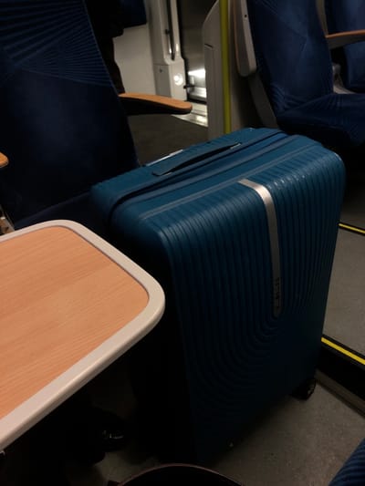 A blue Samsonite suitcase sits atop a train seat, partially obscuring a wooden tray table with a white border. The train interior is visible in the background, including blue patterned seats and a glimpse of the train's window and wall. A hint of a black object, possibly a bag, is on the floor. The image is taken from an elevated angle, looking down towards the suitcase and seating area.