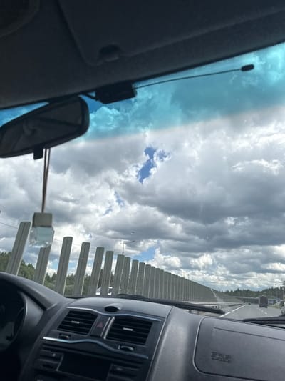 The image shows a view from inside a car, likely on a highway or bridge. The vehicle is moving, as indicated by the blurred scenery. The dashboard and rearview mirror are visible within the car. Outside, the sky is cloudy with patches of blue. A concrete barrier lines the road. Beyond the barrier, trees are visible. Based on the location provided, the image was likely taken in the area near Nagornaya, Russia.