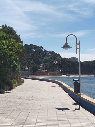 The image depicts a seaside promenade in Rotes, Spain, under a bright, sunny sky with scattered clouds. The promenade curves gently along the coastline, paved with rectangular, light-colored stone tiles. A low concrete wall, painted blue and beige, separates the promenade from the sea. The water appears calm and blue. A series of vintage-style street lamps line the promenade, casting shadows on the tiled surface. A dark gray trash receptacle stands next to one of the lamps. A retaining wall with green foliage climbs the hillside to the left. Beyond the wall, the hillside is covered in trees and dotted with houses and buildings.