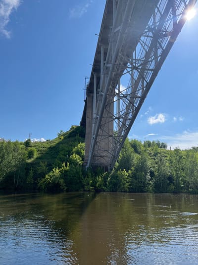 The image shows a view from a body of water, likely a river, looking up at a large, elevated bridge with a complex metal structure. The bridge arches over a green, tree-covered hill. The sky is blue with a few scattered white clouds. Sunlight is visible in the upper right corner, casting lens flare onto the image. The water reflects the sky and the bridge, with ripples on its surface. In the background, on the top of the hill, there appears to be some infrastructure, possibly telecommunication towers. The vegetation is lush and green, indicating a temperate season. There are no people or animals visible in the image. The scene suggests an outdoor, natural setting enhanced by man-made infrastructure, characteristic of a city with both urban development and natural landscapes, such as Kamensk-Uralsky, Russia, known for its industrial heritage and surrounding nature.