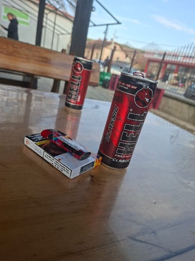 The scene depicts an outdoor setting in Krivodol, Bulgaria, focusing on a wooden table. On the table, from left to right, rests a white pack of cigarettes, atop which a red and black disposable lighter is placed. To the right of these items are two red and black "HELL Classic Energy Drink" cans; the one in the foreground is partially open with its tab lifted, while the one further back appears closed. In the background, behind the table and objects, a wooden bench is visible. To the far left background, a solitary individual in a dark jacket stands, appearing to look down, possibly at a mobile device. Metal fencing or railings run across the mid-ground, and parts of a building with a light-colored facade are visible further back. The environment is illuminated by natural light under a clear blue sky with minimal cloud cover, consistent with an urban or semi-urban outdoor cafe or public seating area.