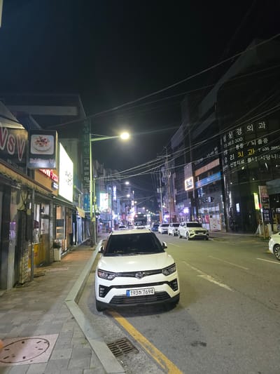 The image depicts a street scene at night in Eumnae-ri, South Korea. A white SUV is parked on the right side of the road, facing forward, with its license plate reading "193하 7694". Several other vehicles are parked or driving on the street. The left side of the street features a sidewalk with shops and businesses. Signs for "VSV", "DAP VSV", and "MIDWAY PIZZA" are visible. A green street lamp illuminates the scene. Buildings line the street, with many featuring illuminated signs in Korean. Power lines crisscross overhead. The overall environment suggests a commercial area at nighttime.