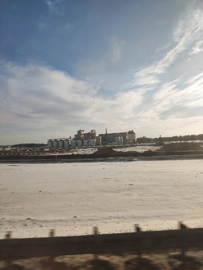 A vast, snow-covered plain stretches to the horizon, leading to an industrial complex. Silos and factory buildings dominate the landscape under a partly cloudy sky. The scene suggests a cold, possibly winter, setting, with the lack of color emphasizing the industrial nature of the area. The bottom edge of the image shows what appears to be a guard rail and ground with debris, possibly indicating this shot was taken from a vehicle or elevated position.
