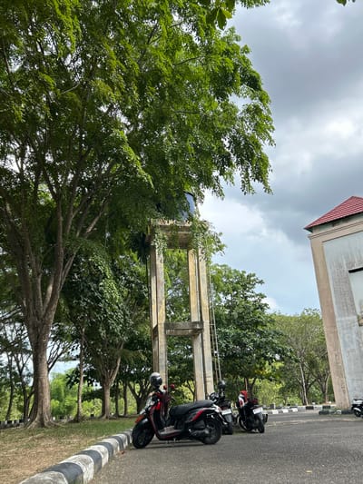 The image depicts an outdoor scene, likely in Reuleut Timu, Indonesia. Dominating the foreground is a large tree with lush green foliage, its branches extending outwards. Behind the tree, a concrete structure, possibly part of a building or elevated walkway, is visible with a red-tiled roof. Several motorcycles are parked in a row along the side of an asphalt road. The road is bordered by a grass verge and a curb with black and white stripes.
The weather appears overcast, with a cloudy sky. The time of day is not precisely discernible but seems to be daytime. The overall atmosphere is calm and still, with no visible human activity besides the parked motorcycles. The presence of motorcycles suggests a location where people might commute or park for a short period, such as near a workplace, educational institution, or recreational area. The vegetation, including the large tree and other trees in the background, indicates a tropical or subtropical environment. No discernible text is visible in the image.