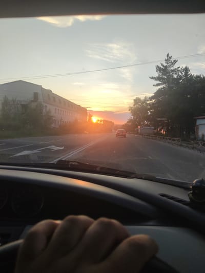 This image, captured from the perspective of a driver inside a vehicle, depicts a scene on a road in Shimanovsk, Russia, during sunset. The sun is low on the horizon, casting a warm, orange glow across the sky and the road, creating lens flare. A car is visible in the distance, driving away from the camera on the right lane of a two-lane road. On the left side of the road, there is a long, white building with a sloped roof, possibly an industrial or commercial structure, bordered by trees and vegetation. A tree is also visible on the right side of the road, with a few buildings and a fence line. The dashboard and the driver's hand on the steering wheel are visible in the foreground, indicating the vantage point of the photograph. The road surface is asphalt, with white lane markings. The sky above is a mix of pale blue and orange hues, with scattered clouds.